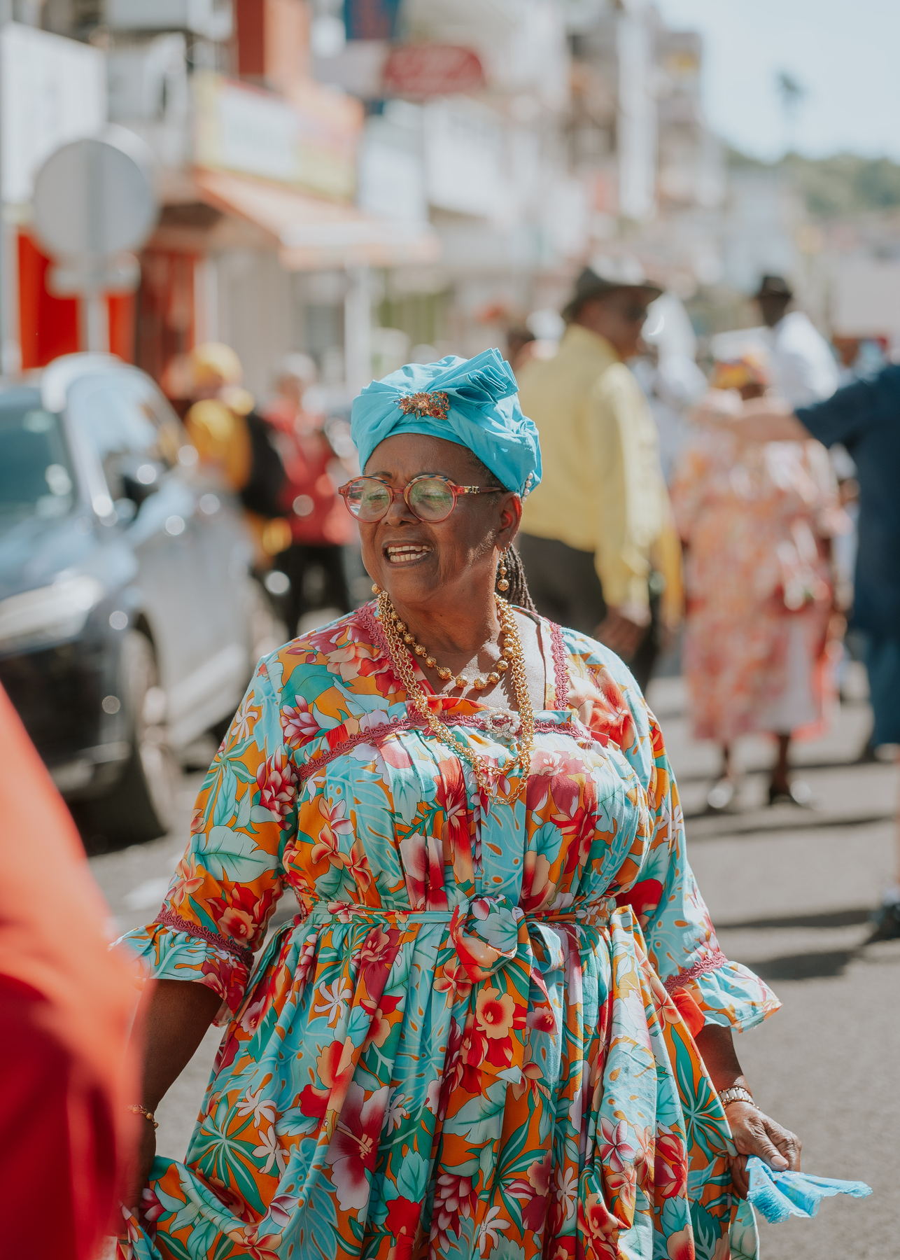 Portrait de rue d'une femme en madras turquoise, façades créoles derrière elle